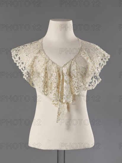 Collar, late 19th century, France. Floral ornaments; women's fashion lace collar with mesh connections and large meshes. Collar sewn with small beads along a polished edge. Lace clothing accessory, width 15 cm, length 230 cm., A mannequin torso in off-white displays a delicate lace collar draped over its shoulders; the lace is a pale cream color with intricate openwork and scalloped edges, forming a ruffled front that hangs in layers and tapers to a pointed end. The mannequin stands against a plain gray backdrop, and the visible support rod beneath it is a darker gray. The overall color palette includes cream, off-white, and shades of gray.