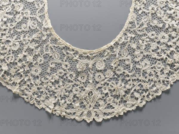 Collar, floral ornaments, fashion, women's fashion, lace, lace collar with mesh connections and large meshes. Collar sewn with small beads from a polished edge. Late 19th century, France. Clothing accessory; lace. Width 15 cm, length 230 cm., A delicate lace collar with an open circular neckline is displayed against a smooth gray background, showing a wide curved shape made of intricate interconnected netting and ornate floral and scroll motifs; the lace appears mainly off-white or cream with subtle variations in tone, with visible fine threads, looped edges, and small raised embroidered details creating a textured, airy pattern across the piece.