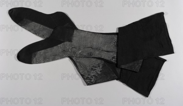Fil d'Ecosse stockings, stockings, fashion, fashion accessories, women's fashion, lingerie, Art Nouveau style. The stockings made of thin, delicate black thread are decorated with openwork machine-made lace on the instep, and the heels and feet are reinforced with double weaving. A ship's head runs from the toes along the entire length of the stockings. ca. 1900, Europe, clothing, length 62 cm., A pair of black sheer stockings laid flat on a light gray background, showing decorative lace-like patterned panels and solid darker toe and heel areas; the fabric displays a mix of transparent mesh and opaque knit with floral and scalloped motifs, and the overall tones in the image are black, dark gray, and light gray.
