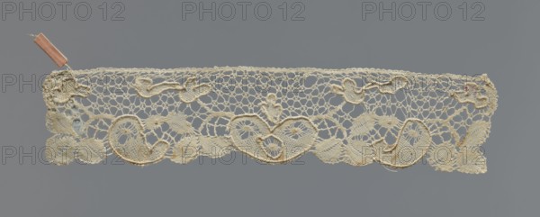 Cuff, cuffs; fashion accessory; lace. Pattern of leaves and palmettes at the lower edge. Mesh background with triangular and round eyelets. Straight upper edge. 2nd half of the 19th century, Germany . Clothing accessory; width 6 cm, length 29 cm., A narrow strip of delicate lace in shades of off-white and cream is set against a solid gray background; the lace has an open, net-like upper section with a decorative lower border featuring a repeating pattern of heart-like swirls and leaf shapes, and a few raised, corded outlines that add texture; small irregularities and slight discolorations in pale brown and tan appear in places along the lace, and a small pinkish ribbon is attached near one end.