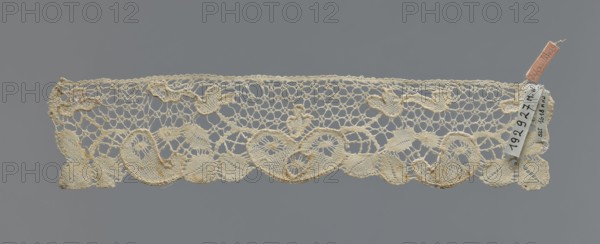 Cuff, repossession provenance; fashion accessory cuff; lace; pattern of leaves and palmettes at the lower edge; mesh background with triangular and round eyelets; straight upper edge; 2nd half of the 19th century; Germany . Dimensions: width 6 cm, length 29 cm., A narrow strip of lace with an openwork pattern laid flat against a neutral gray background, showing a scalloped lower edge with curved, leaflike and teardrop motifs and an upper edge with a more irregular honeycomb-like mesh; the lace appears off-white to cream with areas of slightly darker beige tones and small accents of pale brown discoloration, and there is a small fabric tag attached at one end in a muted pinkish color with a small pale cloth label nearby.