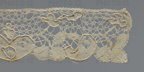 Cuff, repossession provenance; cuffs; fashion accessory; lace; pattern of leaves and palmettes at the lower edge; mesh background with triangular and round eyelets; straight upper edge; 2nd half of the 19th century; Germany probable place of origin; clothing accessory; width 6 cm; length 29 cm., A section of openwork lace framed against a gray background, showing a delicate network of interconnected loops and mesh in an off-white to cream color with areas of slightly darker beige discoloration and a small spot of pale pink; the lace features several rounded and scalloped motifs with raised corded outlines and densely stitched inner areas, a few leaf-like shapes and floral-inspired patterns, and some loose and frayed thread ends visible along the lower edge.