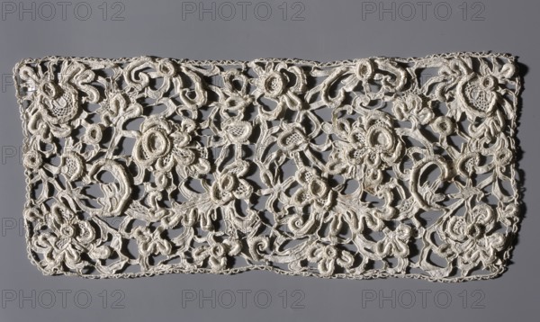 Lace fragment, Plant ornaments and plant tendril; contours of flowers and leaves executed in relief. Almost no joints. Straight edges finished with serrations and picots. Imitation of 17th-century Venetian lace. Probably a cuff. 2nd half of the 17th century, Venice. Lace, height 36 cm, width 15 cm., A rectangular piece of openwork lace with an intricate floral and scroll pattern laid flat on a smooth background; the lace is an off-white or cream color with raised embroidered outlines and textured fill areas forming a dense network of flowers, leaves, loops, and curving stems, while the background is a uniform gray that contrasts with the lighter lace.