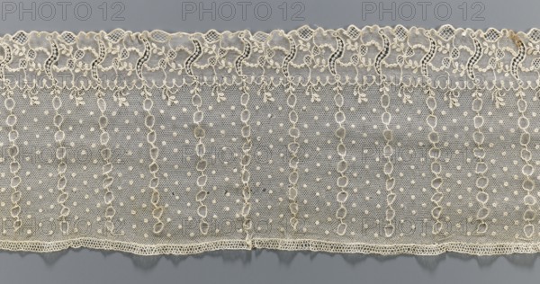 Lace fragment, Alençon lace, lace; plant ornaments. At the bottom, an edging ornament running in a clearly separated strip, with plant motifs and an openwork form resembling the letter S repeated regularly. Higher on the mesh, oval stripes between which projections: small diameter points. 1st half of the 18th century, France. Material: fabric. Width 17 cm, length 206 cm., A horizontal strip of delicate lace fabric in a pale cream color lies against a muted gray background, showing an upper scalloped edge with ornate looping floral motifs and a lower straight edge with a finer scalloped trim; the main body of the lace displays a repeating vertical pattern of open oval chain-like columns separated by mesh areas dotted with small raised bobbles, and the overall texture appears lightweight and finely netted with subtle variations in thread thickness and tiny decorative loops along the borders.