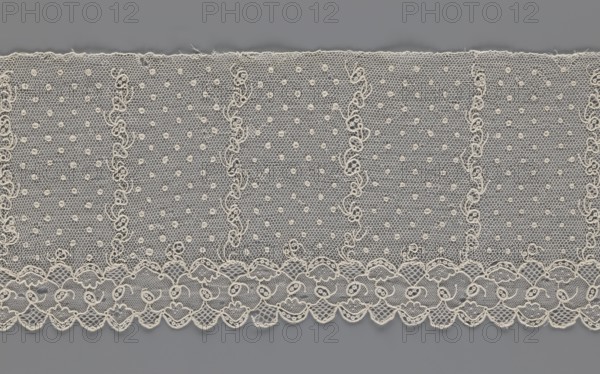 Lace fragment, A strip with a serrated edge on one side and an edge-type ornament on the serrated side; above this are narrow strips of stylized plant ornament with an onion-shaped flower motif and, between them, small round dots in a regular pattern against a mesh background. 1st half of the 19th century, Europe. Materials: lace, fabric. Dimensions: width 12.5 cm, length 210 cm., A length of delicate lace fabric with an overall off-white or cream color laid flat against a gray background, featuring a repeating pattern of fine hexagonal netting filled with small circular dots and vertical wavy embroidered lines, and a scalloped lower edge adorned with ornate loops and floral-like motifs; the lace shows subtle texture variations and tiny loose threads along the top edge, with visible contrast between the cream lace and the mid-gray background.