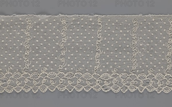 Lace fragment, strip with a serrated edge on one side and an edge-type ornament on the serrated side; above this are narrow strips of stylized plant ornament with an onion-shaped flower motif and, between them, small round dots in a regular pattern against a mesh background. 1st half of the 19th century, Europe. Lace, fabric; width 12.5 cm, length 210 cm., A wide piece of delicate lace fabric with a repeating ornate pattern, shown against a flat background; the lace is light ivory or cream in color with a fine hexagonal mesh and small raised circular dots scattered throughout, vertical wavy embroidered lines creating panels, and a scalloped lower edge featuring floral-like motifs and looping embroidered details; the background visible through the lace and at the top and bottom is a smooth mid-gray.