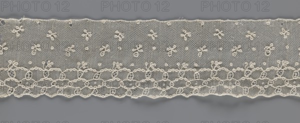 Lace fragment, edge ornament on one side — a kind of net with numerous loops small, round and slightly larger oval into which small twigs are woven. Above the edge ornament, against a netting background, in regular projections alternate: a twig and a round point. 18th–19th century, France. Materials: lace, fabric. Dimensions: width 8.5 cm, length 408 cm., A horizontal strip of delicate lace in an off-white or cream color stretches across a flat gray background, featuring a fine hexagonal mesh ground dotted with small embroidered floral motifs and tiny round accents; the lower edge has a scalloped border composed of interconnected loops and oval shapes with denser embroidery, while the upper edge is relatively straight with slight waviness and occasional small embroidered loops along it; the overall pattern is airy and detailed, with visible variations in thread thickness and subtle shadows where the lace lifts from the background.