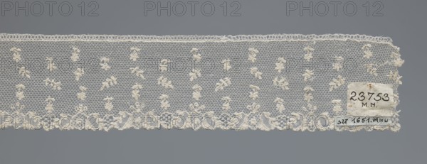 Fragment of lace, one-sided strip of edge ornament made of plant forms with a motif of a pinnately leafed twig; above the edge ornament, against a mesh background in projections, an alternating twig with a single flower and a pinnately leafed twig. Lace, 18th century, France. Width 6 cm, length 63 cm., ., A horizontal length of delicate lace displayed against a neutral gray background, showing a fine hexagonal net ground with a repeated motif of small clustered floral-like knots in an off-white or cream color, a scalloped lower edge with denser embroidered floral and scroll designs, a straight upper edge with a narrow looped border, and a small rectangular patch of fabric attached near one end in similar cream tones.