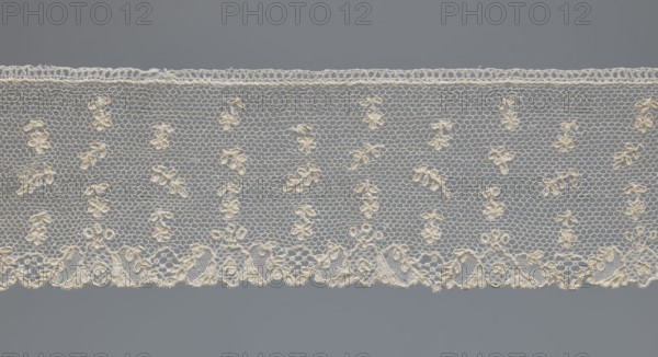 Fragment of lace, one-sided strip of edge ornament made of plant forms with a motif of a pinnately leafed twig; above the edge ornament, against a mesh background, alternating projections of a twig with a single flower and a pinnately leafed twig. Lace, 18th century, France. Width 6 cm, length 63 cm., A horizontal strip of delicate lace with a scalloped lower edge and a straight upper edge, featuring a repeating floral motif worked into a fine hexagonal net ground; the lace appears in shades of off-white and cream set against a medium gray background, with the floral motifs and decorative border showing slightly warmer cream tones while the netting reads as pale off-white.