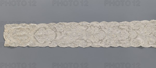 Lace fragment, Binche lace, plant ornaments, floral ornaments; floral and plant pattern in a candelabra arrangement, with flowers enclosed in an oval at the bottom and freely drawn at the top; mesh background of the ißsgrond and point de neige types; ca. 1750; Binche; lace; width 10 cm; length 118 cm, A long horizontal strip of delicate lace in off-white and cream tones lies against a smooth gray background, featuring an intricate floral and foliate pattern with scalloped edges; the lace shows a repeating design of large stylized blossoms, leaves, and curling vines rendered in fine openwork and denser embroidered areas, with a few areas of slightly darker discoloration and subtle shadowing that emphasize the texture and raised threadwork.