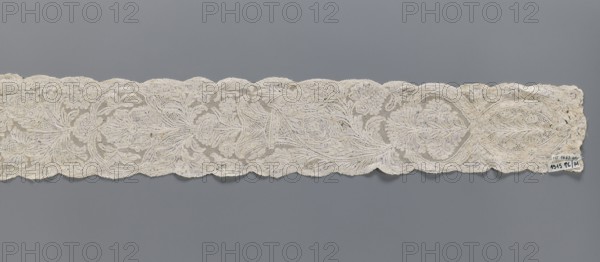 Lace fragment, Binche lace, plant ornaments, floral ornaments. Floral and plant pattern in a candelabra arrangement, with flowers enclosed in an oval at the bottom and freely drawn at the top. Mesh background of the ißsgrond and point de neige types. Ca. 1750. Lace, width 10 cm, length 118 cm., A long narrow piece of lace with a scalloped top and bottom edge stretches across the image against a solid gray background, showing an intricate floral pattern with flowing vine-like motifs and open mesh areas; the lace appears in shades of off-white, cream, and light beige with subtle variations in tone, and there is a small irregularity at one end where the fabric looks slightly frayed.