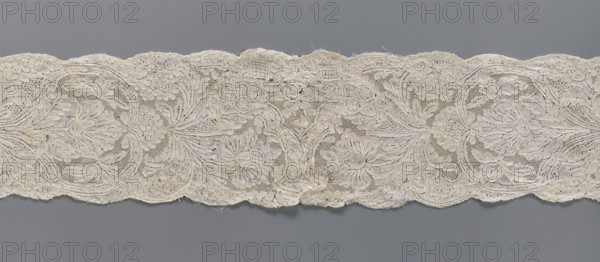 Lace fragment, Binche lace, plant ornaments, floral ornaments, floral and plant pattern in a candelabra arrangement, with flowers enclosed in an oval at the bottom and freely drawn at the top. Mesh background of the ißsgrond and point de neige types, ca. 1750, Binche; lace, width 10 cm, length 118 cm., A long scalloped band of lace in off-white and cream tones lies horizontally against a muted gray background, showing an ornate floral pattern with several large stylized blooms, leaves, and curving vines worked in fine thread; the lace has areas of open mesh and denser embroidered motifs, with a few frayed edges and small irregularities in the fabric texture visible along the scalloped borders.