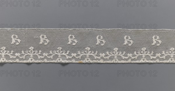 Lace fragment, plant ornaments, Louis XVI style, Malines Mechelen lace. At the lower edge there are two trails of fine tendrils, and between them a net with issgrond, mesh. Above, a net with drochelle mesh, on it projections of leafy branches. Contours of motifs reinforced with thread. Late 18th century. Lace; width 7.5 cm, length 146 cm., A horizontal strip of delicate lace in off-white sits against a smooth gray background, featuring a fine hexagonal mesh with a scalloped floral border along one edge and a repeating motif of small floral-like embroidered shapes spaced above the border, showing tiny loops and raised thread details and a slight darker discoloration on the lower edge; visible colors are off-white, gray, and a small patch of tan.