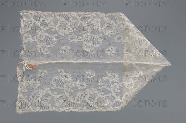 Fragment of lace, lace. Inside the motifs there are varied mesh backgrounds: drochel, ißsgrond, and valanciennes. At the top there is a lille type mesh. Background: fond chant, point de Tulle, point de Drochelle. 2nd half of the 18th century, Mechelen. Lace, width 13.5 cm, length 64 cm., A triangular piece of delicate lace with floral and scrollwork patterns in off-white and cream tones lies flat against a smooth gray background, showing a mesh netting structure and scalloped edges; a small fabric tag in muted brown and pale beige is attached near one corner.
