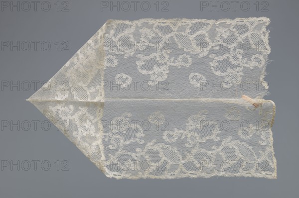 Fragment of lace by, lace with varied mesh backgrounds—drochel, ißsgrond, and valanciennes; top with a lille type mesh; background: fond chant, point de Tulle, point de Drochelle; 2nd half of the 18th century; Mechelen; lace; width 13.5 cm, length 64 cm, A triangular piece of delicate lace fabric displayed against a gray background, the lace is a pale off-white or cream color with an open netting structure and floral and scrolling motifs woven throughout; the fabric shows a fold forming a pointed tip on one side and a longer rectangular section extending across, with frayed edges and a small area of slight discoloration in a pale tan or peach tone near one corner, and the overall appearance includes a few fine loose threads and subtle variations in the cream shading.