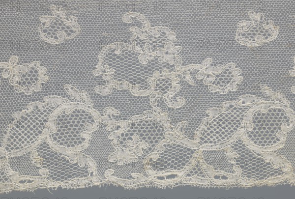 Fragment of lace, lace. Inside the motifs there are varied mesh backgrounds: drochel, ißsgrond, and valanciennes. At the top there is a lille type mesh. Background: fond chant, point de Tulle, point de Drochelle. 2nd half of the 18th century, Mechelen. Width 13.5 cm, length 64 cm., A delicate lace fabric fills the frame with an open hexagonal mesh background and ornate floral and scroll motifs worked in a slightly thicker thread; the lace is predominantly off-white or cream against a muted gray background, showing a few irregular edges and tiny loose fibers along the bottom, and the overall pattern includes several rounded shapes and looping decorative elements creating a repeating, airy design.
