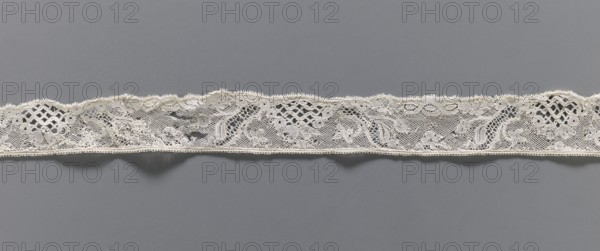 Lace fragment, floral ornaments, Malines lace, pattern with motifs of winding ribbons, flowers, and rococo elements; Point de drochelle background. 2nd half of the 18th century, Mechelen. Lace, width 3.5 cm, length 83 cm., A narrow strip of delicate lace runs horizontally across a flat gray background, showing an intricate openwork pattern with floral and leaf motifs, scalloped upper edge and a straighter lower edge, featuring areas of fine netting, looped decorative borders, and denser embroidered sections; the lace is primarily off-white or cream against a medium gray backdrop.
