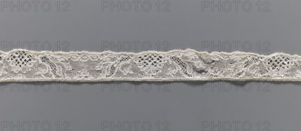 Lace fragment, Floral ornaments. Malines lace. Pattern with motifs of winding ribbons, flowers, and rococo elements. Point de drochelle background. 2nd half of the 18th century. Mechelen. Lace, width 3.5 cm, length 83 cm., A horizontal strip of delicate lace with floral and geometric openwork motifs lies across a smooth background; the lace is an off-white or cream color with intricate netted areas and scalloped top edge, and the background is a uniform medium gray.