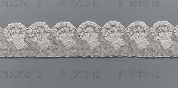 Fragment of lace, Malines Mechelen lace, 18th century, from Henryków Heinrichau, lace, width 6 cm, length 62 cm, A strip of delicate lace with a scalloped top edge lies horizontally against a uniform gray background; the lace is off-white and features a repeating pattern of ornate floral bouquets worked into a fine hexagonal mesh, with the scallops forming rounded arches above each bouquet and a straight lower edge below; the floral motifs show clustered rounded shapes and textured stems, and the lace displays subtle variations in thread density and tiny openings where the mesh is visible.