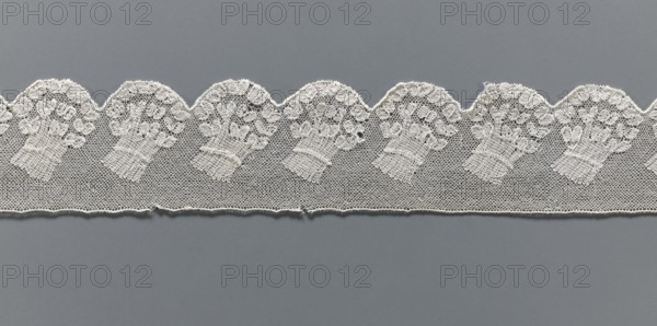 Fragment of lace, Malines Mechelen lace, 18th century, Henryków German: Heinrichau, lace, width 6 cm, length 62 cm., A horizontal strip of delicate white lace with a scalloped upper edge rests against a muted gray background; the lace features a repeating floral bouquet motif worked in fine thread over a hexagonal net ground, showing a row of scallops with ornate flower clusters and textured stems, and the lower edge of the lace has a subtle finished border with a few small loose threads. The visible colors are white in the lace and gray in the background.