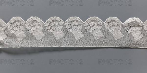 Fragment of lace, repossession provenance noted, Malines Mechelen lace, 18th century, Henryków Heinrichau — museum repository, lace, width 6 cm, length 33 cm, A scalloped strip of lace in white stretches horizontally across a smooth gray background, featuring a repeating floral bouquet motif worked into a fine netted pattern; the lace shows delicate embroidered textures, curved scallop edges, and areas where the netting and denser embroidery create slight variations in tone, with visible white threads and shadows against the gray surface.