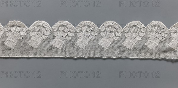 Fragment of lace, Malines Mechelen lace, 18th century, Henryków Heinrichau, lace, width 6 cm, length 33 cm, A strip of delicate white lace with a scalloped upper edge lies horizontally across a flat gray background, the lace featuring a repeating motif of bouquet-like floral shapes worked into a fine netted mesh; the visible colors are white and gray.