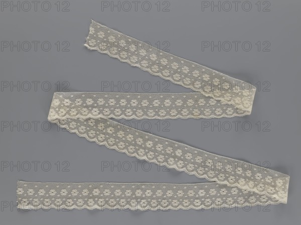 Lace fragment, tied in the form of a belt with one-sided arched serrations. Regular ornament: rosettes and points arranged in rows. Mid-19th century, possibly Flanders. Lace, width 7 cm, length 223.5 cm., A length of delicate lace ribbon arranged in a loose zigzag across a flat surface, showing a repeating floral motif within a fine mesh, scalloped edges, and slight irregularities at the cut ends; the lace is an off-white or cream color set against a smooth medium-gray background.