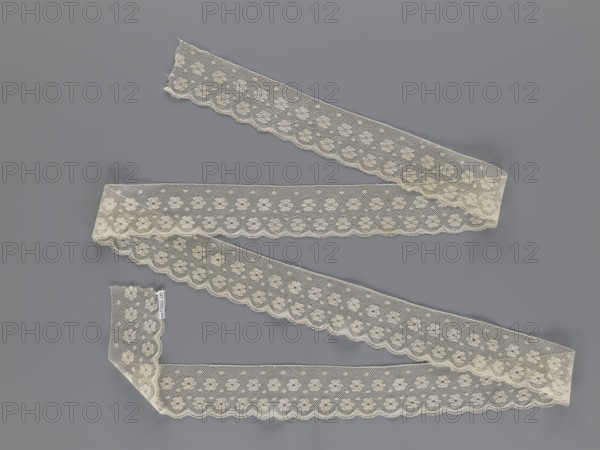 Lace fragment, tied in the form of a belt with one-sided arched serrations. Regular ornament: rosettes and points arranged in rows. Mid-19th century, possibly Flanders. Lace, width 7 cm, length 223.5 cm. Maker: Kublicki Piottuch, Alfred 1861–1945., A length of scalloped lace ribbon arranged in a zigzag pattern on a flat surface, showing a delicate floral mesh design with a scalloped edge; the lace is an off-white or cream color and the background is gray.