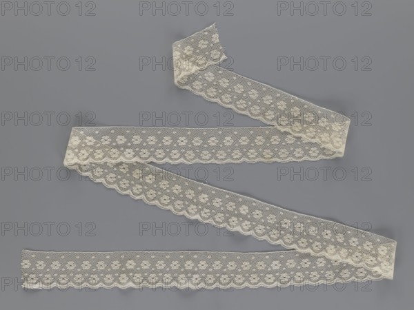 Lace fragment tied in the form of a belt with one-sided arched serrations. Regular ornament of rosettes and points arranged in rows. Mid-19th century, possibly Flanders. Lace, width 7 cm, length 223.5 cm., A length of delicate lace ribbon arranged in a zigzag pattern on a flat surface, the lace showing a repeating floral motif and scalloped edges, with visible net-like openwork and slight fraying at some ends; the lace appears off-white or cream, and the background is a muted gray.