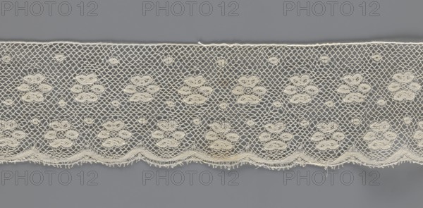 Lace fragment, tied in the form of a belt with one-sided arched serrations. Regular ornament: rosettes and points arranged in rows. Mid-19th century, possibly Flanders. Lace, width 7 cm, length 223.5 cm. Creator: unknown., A horizontal strip of delicate lace with a repeating floral motif set against a flat background, showing a net-like openwork of diamond-shaped holes and clusters of rounded, petal-like embroidered elements; the lace has a decorative scalloped lower edge with tiny looped fringes and an upper edge finished with a straight selvage; visible colors include off-white or cream for the lace and a muted gray for the background.