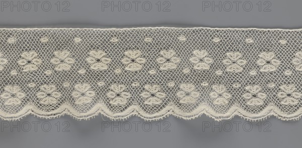 Lace fragment, tied in the form of a belt with one-sided arched serrations. Regular ornament: rosettes and points arranged in rows. Mid-19th century, possibly Flanders. Lace, width 7 cm, length 223.5 cm., A horizontal strip of delicate lace in off-white with a repeating floral motif set against a gray background, featuring a net-like mesh between the floral clusters, small circular lace accents scattered throughout, and a scalloped lower edge with textured fringe; colors visible include off-white and gray.