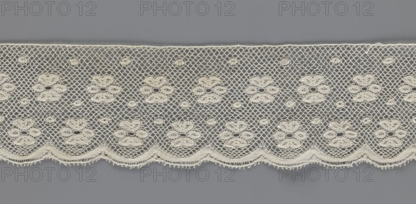 Lace fragment, tied in the form of a belt with one-sided arched serrations. Regular ornament: rosettes and points arranged in rows. Mid-19th century, possibly Flanders. Lace; width 7 cm, length 223.5 cm. Maker: Kublicki Piottuch, Alfred 1861–1945., A wide horizontal strip of lace in off-white is laid against a flat gray background, showing a repeating floral-inspired openwork pattern with a network of diamond-shaped mesh; the lace features a scalloped lower edge with small looped picot details and a top straight edge, and within the mesh there are several clustered oval motifs forming flower-like shapes and smaller circular eyelet motifs dispersed throughout, all in off-white contrasting with the gray.
