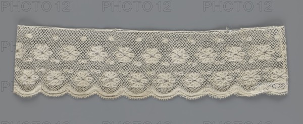 Fragment of lace, lace, a section of a belt sewn into a closed circle with one-sided arched serrations. Regular ornament: rosettes and points arranged in rows. Mid-19th century, Flanders, lace, width 7 cm, length 50 cm., A rectangular piece of openwork fabric with a repeating floral lace pattern in off-white cream color set against a solid gray background; the lace has a diamond mesh ground with a few larger scalloped floral motifs arranged in rows and a scalloped lower edge, with delicate looped fringe along the scallops and some irregular threads visible along the top edge.