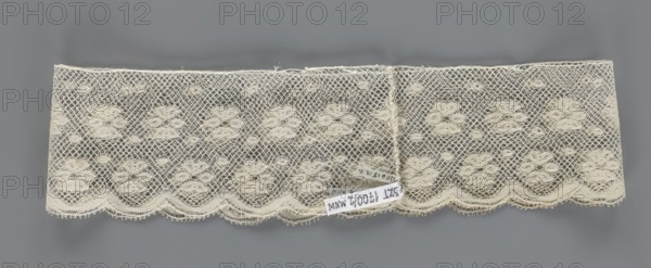 Fragment of lace, a section of a belt sewn into a closed circle with one-sided arched serrations. Regular ornament: rosettes and points arranged in rows. Mid-19th century, Flanders, lace, width 7 cm, length 50 cm., A rectangular piece of delicate lace with an open netted pattern and floral motifs, laid flat against a gray background; the lace is a creamy off-white color with a scalloped lower edge and a few small loose threads along the top; the netting forms diamond-shaped holes surrounding clusters of rounded floral shapes, and there is a small group of labels attached near the lower center that contrast with the lace.