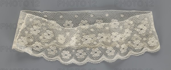 Fragment of lace, lace. A section of a belt sewn into a closed circle with one-sided arched serrations. Regular ornament: rosettes and points arranged in rows. Mid-19th century, possibly Flanders. Lace, width 7 cm, length 50 cm., A length of delicate lace fabric displayed against a neutral background, showing an open mesh pattern with a repeating floral motif and scalloped edge; the lace appears in off-white or cream tones while the background is a medium gray, and the fabric has slight folds and shadowing that create areas of lighter and darker cream.