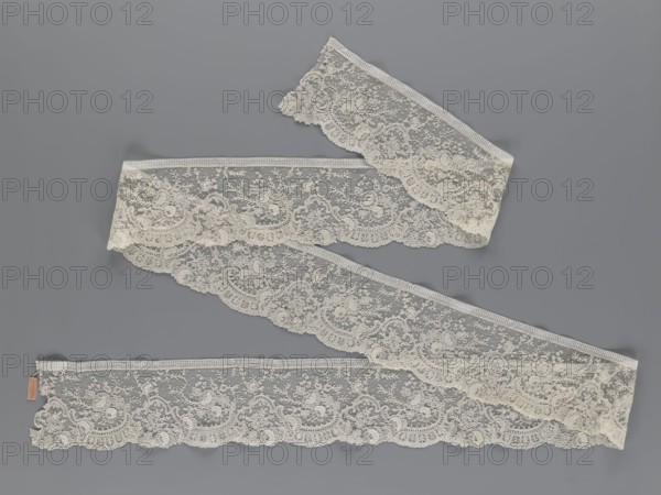 Lace fragment, lace, floral ornaments with rocaille motifs, over-stylized fan-shaped ribbons with arcades, twigs, dotted background, point de gaze ground. Lower edge serrated to match motifs; upper edge straight with a bobbin insert. Some rose flowers are in relief with petals made separately and attached by one edge. 1st half of the 19th century, France, lace, width 15 cm, length 298 cm, A length of delicate lace ribbon arranged in gentle S-shaped curves against a smooth background, the lace displaying an intricate floral and leafy pattern with scalloped edges and fine netting, the visible colors being off-white for the lace and a muted gray for the background.