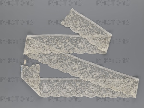 Lace fragment, lace, floral ornaments with rocaille motifs, over-stylized fan-shaped ribbons with arcades, and twigs; dots in the background; point de gaze background. Lower edge serrated to match motifs; upper edge straight with a bobbin insert. Some rose flowers worked in relief, with petals made separately and attached by one edge. 1st half of the 19th century, France, lace; width 15 cm, length 298 cm., A length of delicate lace fabric arranged in a loose S-shaped layout against a flat background, showing an intricate floral and scrollwork pattern with scalloped edges; the lace appears in off-white or cream tones with subtle variations in shading, and the background is a muted cool gray; a small fabric tag is attached near one end of the lace.