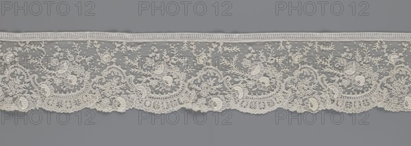 Lace fragment, lace, floral ornaments with rocaille motifs, over-stylized fan-shaped ribbons with arcades, and twigs; dots in the background; point de gaze background; lower edge serrated to match motifs, upper edge straight with a bobbin insert; some rose flowers made in relief with petals made separately and attached by one edge; 1st half of the 19th century; France, lace; width 15 cm, length 298 cm., A horizontal strip of lace with an intricate floral and leafy pattern in off-white against a medium gray background, showing a scalloped lower edge and a straight upper border; the lace features a mix of solid embroidered motifs and delicate openwork, with several clusters of stylized flowers, curling vines, small dot-like accents, and leafy sprigs repeated across the length, and ornamental curved arches and fan-like shapes integrated into the lower scalloped section.