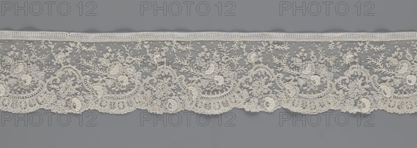 Lace fragment, Lace with floral ornaments, rocaille motifs, over-stylized fan-shaped ribbons with arcades, twigs, and dotted background. Point de gaze ground. Lower edge serrated to match motifs; upper edge straight with a bobbin insert. Some rose flowers are in relief, with petals made separately and attached by one edge. 1st half of the 19th century, France, lace; width 15 cm, length 298 cm., A horizontal strip of decorative lace in cream color set against a gray background, featuring an intricate floral and botanical pattern with a few scalloped edges along the bottom, several repeating arched motifs, and a fine dotted mesh filling between the embroidered elements; the lace shows detailed leaf and flower shapes, curved vine-like lines, and a narrow woven border along the top.
