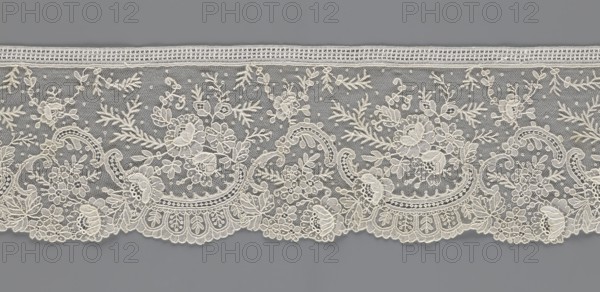 Lace fragment, lace with floral ornaments including rose flowers—some modeled in relief with petals separately made and attached by one edge. Features rocaille motifs, over-stylized fan-shaped ribbons with arcades containing twigs, a point de gaze background with dotted background. Lower edge serrated to match motifs; upper edge straight with a bobbin insert. 1st half of the 19th century, France, lace, width 15 cm, length 298 cm., A length of delicate lace displayed horizontally against a solid background, featuring an off-white or cream floral embroidery on a fine netting; the lace shows a scalloped lower edge with ornate rounded motifs and clustered floral shapes, leafy stems, and vine-like scrolls rising into a banded top edge with a narrow decorative trim; the netting background appears slightly darker than the embroidery, creating contrast between the cream lace and a muted gray backdrop.