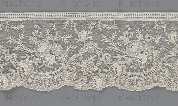 Lace fragment, lace, floral ornaments with rocaille motifs, over-stylized fan-shaped ribbons with arcades, twigs, dotted background, point de gaze background. Lower edge serrated to match motifs; upper edge straight with a bobbin insert. Some rose flowers formed in relief with petals separately made and attached by one edge. 1st half of the 19th century, France, width 15 cm, length 298 cm., The image shows a length of delicate lace with a floral and scroll pattern against a gray background; the lace is ivory or off-white and features a fine net ground dotted with small picot-like points, a scalloped lower edge with ornate motifs, and an upper straight edge with a narrow band; the design includes a group of stylized flowers, leaves, buds, and curling vines arranged in repeating panels, with varying stitch textures creating raised and open areas; visible colors are gray for the background and ivory or off-white for the lace.