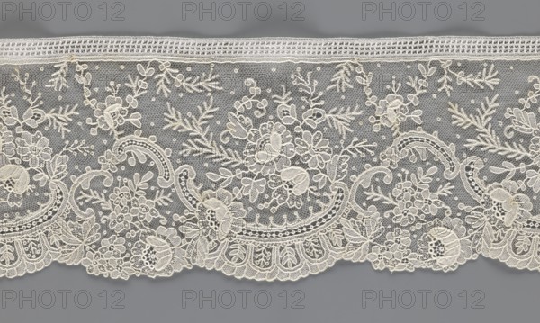 Lace fragment, lace with floral ornaments, rocaille motifs, over-stylized fan-shaped ribbons with arcades, and twigs; dots in the background; point de gaze background; lower edge serrated to match motifs, upper edge straight with a bobbin insert; some rose flowers worked in relief with petals made separately and attached by one edge; 1st half of the 19th century; France probable; lace; width 15 cm, length 298 cm., A length of delicate lace with an intricate floral and foliate pattern laid against a neutral background, showing a scalloped lower edge and a straight upper border; the lace features a fine hexagonal mesh filled with a group of embroidered flowers, leaves, scrolls, and small dot motifs, with emphasized outlines and textured fill stitches creating raised details; visible colors include off-white or cream embroidery atop a medium gray background, with subtle variations in the cream threads giving a slightly warm tone against the cool gray.