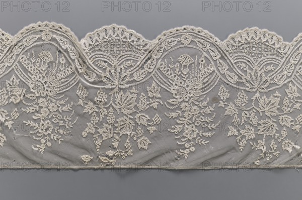 Lace fragment, bobbin lace, mid-19th century, Brussels. Two ribbons: one wider with leaves, the other narrower with a chain; interlacing to create ovals containing garlands. Branches with flowers or vine branches with fruit extend upward from the ovals. Background in fond de gaze. Lower edge semicircularly serrated, upper edge straight. Materials: lace. Width 28 cm, length 187 cm., A scalloped piece of lacework with an intricate floral and leaf embroidered pattern laid against a smooth background; the lace is a creamy off-white with detailed loops, mesh netting, and raised stitched motifs forming bouquets, sprigs, and clustered blossoms, while the background is a muted gray that contrasts with the lace; the lace edge shows repeating rounded arches with decorative openwork and stitched borders, and the lower edge finishes with a narrow stitched hem.