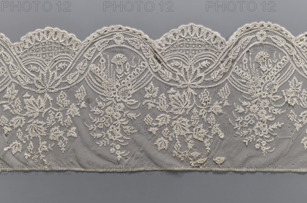 Lace fragment, bobbin lace, mid-19th century, Brussels. Plant ornaments; one ribbon is wider with leaves, the other narrower with a chain. Interlacing ribbons create ovals containing garlands; branches with flowers or vine branches with fruit extend upwards from the ovals. Background is fond de gaze. Lower edge semicircularly serrated, upper edge straight. Width 28 cm, length 187 cm., A decorative fabric panel with an ornate scalloped top edge showing a repeating curved motif, covered in intricate embroidered floral and leaf patterns; the embroidery is a light cream color set against a muted gray background, with delicate netting visible beneath the raised stitched designs; the floral motifs include clustered buds, stylized blossoms, and trailing vines arranged in a repeating layout across the panel; the scalloped border features denser stitching with geometric and looped details, while the lower field shows more open net areas and scattered embroidered sprays; overall colors visible a...