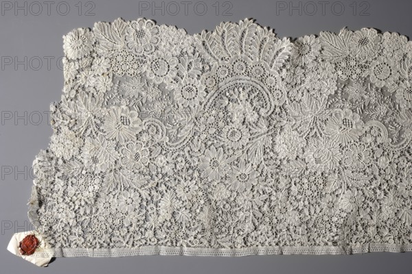 Fragment of Brussels lace; floral ornaments of densely placed roses, leaves, rocaille and over-stylized rooster combs. The flowers and bridge background were made using the bobbin technique. Some sections were executed on a point de gaze background, both background and flowers using needlework technique. The background contains only sparse bridge connections. The central section features irregular medallions on a point de gaze background with small flowers. Lace, ca. 1850, Belgium. Length 500 cm, width 34 cm., A wide piece of lace fabric with intricate floral and leaf patterns spread against a solid background, showing delicate openwork, circular flower motifs, scalloped edges, and dense embroidered areas; the lace appears in an off-white or cream color, the background is gray, and a small folded paper corner with a red wax seal is visible at one edge.