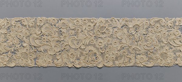 Fragment of lace, lace, plant ornaments, ca. 1700, France, lace, width 17 cm, length 157 cm, A long strip of intricately patterned beige lace laid horizontally against a smooth gray background, featuring a dense floral and foliate design with a few large stylized blossoms, several smaller rosette-like motifs, and many curving paisley-shaped elements; the lace shows varied openwork areas and raised embroidered outlines, with visible texture from delicate netting, looping threads, and tiny picot edges, and the visible colors are beige and gray.