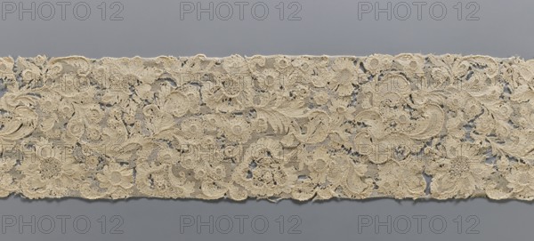 Fragment of lace, plant ornaments, ca. 1700, France, lace, width 17 cm, length 157 cm, A horizontal strip of intricate lace with floral and leafy motifs stretches across a plain background; the lace appears in shades of beige and off-white with textured openwork, showing a variety of scalloped edges, circular flower shapes, curling tendrils, and densely stitched areas interspersed with more transparent mesh, all set against a smooth grey backdrop.