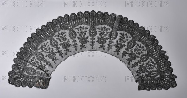 Collar, floral ornaments, Chantilly lace, lace, clothing accessory, lace collar, Regular, reported geometric-floral pattern with divisions marked by arcs and loops of a belt running along the circumference, with floral sprigs in the divisions; tulip cups on the outer edge. 2nd half of the 19th century, France. Width 12 cm, length 73 cm., A semi-circular piece of lace with an ornate scalloped edge spread across a light background, showing a repeating floral and leafy pattern worked into fine netting; the lace displays dense decorative motifs along the outer scallops and more open netting toward the inner curve, with visible layered textures and small circular openings in the trim, and the colors visible are shades of black and gray against a pale gray background.