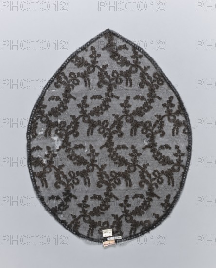 Lace, plant ornaments, in the shape of an oval pointed on one side. A narrow strip of geometric ornamentation on the outer edge. The field is filled with leafy and floral tendrils forming a kind of net fond de Tulle. 19th century, France; fabrics; height 46 cm, width 35 cm., An oval-shaped lace fabric piece with a scalloped dark edging and an all-over floral vine pattern in dark brown on a light gray sheer background, placed on a pale gray surface; a small cluster of light-colored rectangular tags is attached near the lower edge.