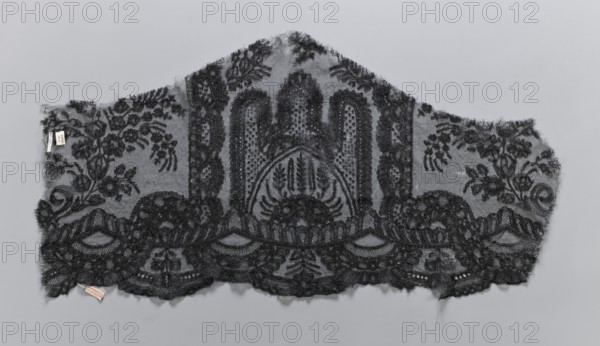 Lace fragment, plant ornaments. Preserved ruffle fragment features a symmetrical ornament of a central arch with three pinnacles. Serrated edge decorated with a wide band of edge ornament composed of arch segments with geometric ornament and an interwoven tendril from a band of plant-geometric ornament. 19th century, France. Lace, width 38 cm, length 74 cm., A scalloped piece of delicate black lace fabric with an ornate floral and foliate pattern laid flat against a neutral gray background; the lace shows a central arched motif with dense embroidered detailing and dotted netting, flanked by symmetrical botanical sprays and scrolling leaves, and a lower scalloped border of dense floral embroidery and openwork; loose threads and small frayed edges are visible along the outer edges, and a small fabric tag is attached near one corner.